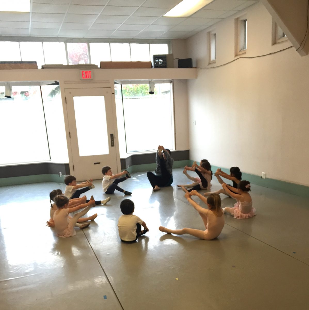 A group of young dancers sit in a circle and stretch with their teacher