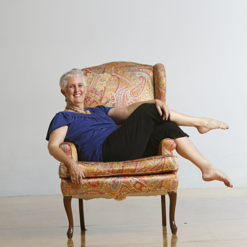 Beth Hoge wears a purple top and black pants. She sits sideways in a chair with legs crossed