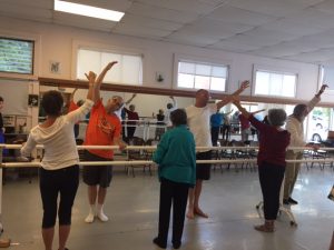 Various dancers in Dance for PD standing at the barre and reaching one arm.