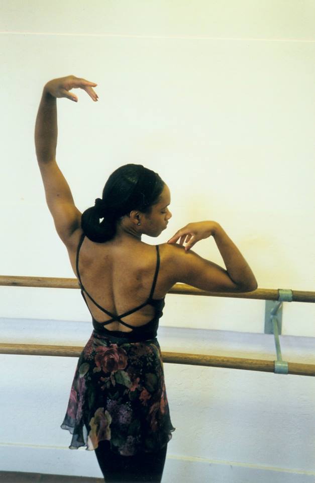 Kimberly faces away from the camera, looking to her right. She wears a black leotard and floral skirt.