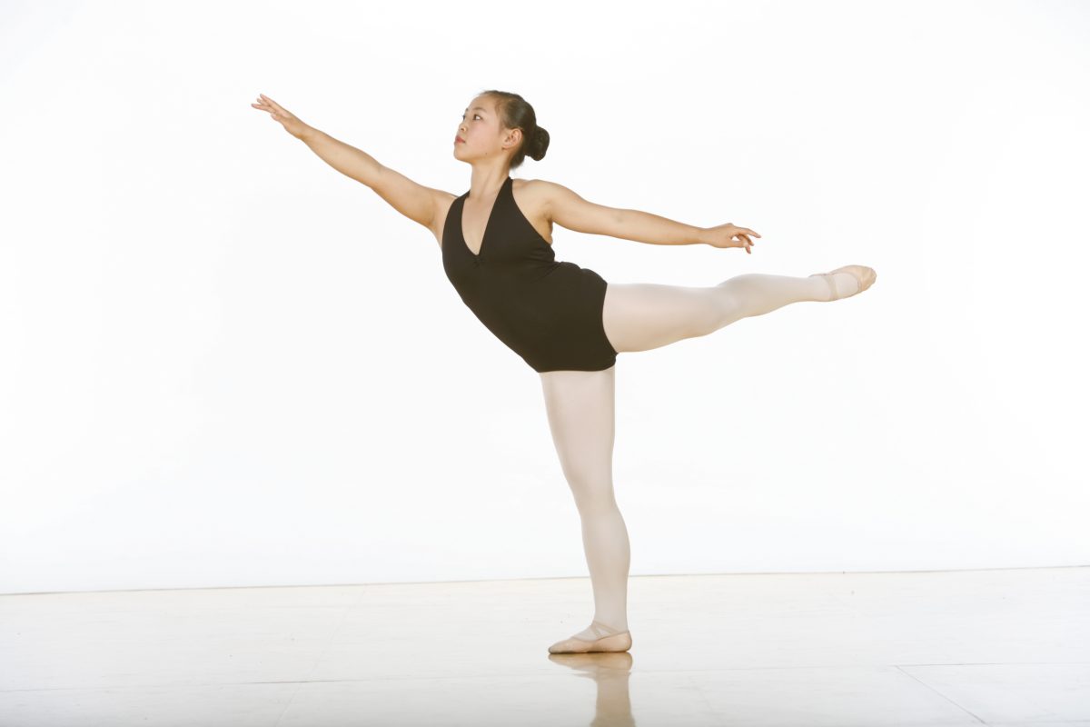 A dancer in a black leotard and pink tights in first arabesque en releve.