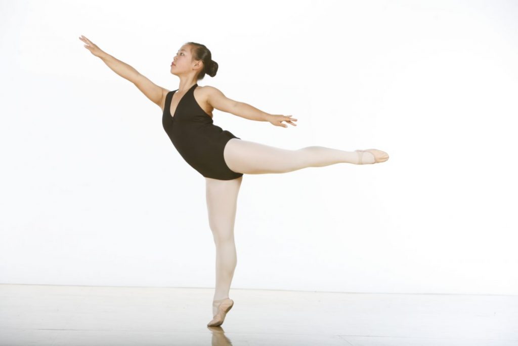 A dancer in a black leotard and pink tights in first arabesque en releve.