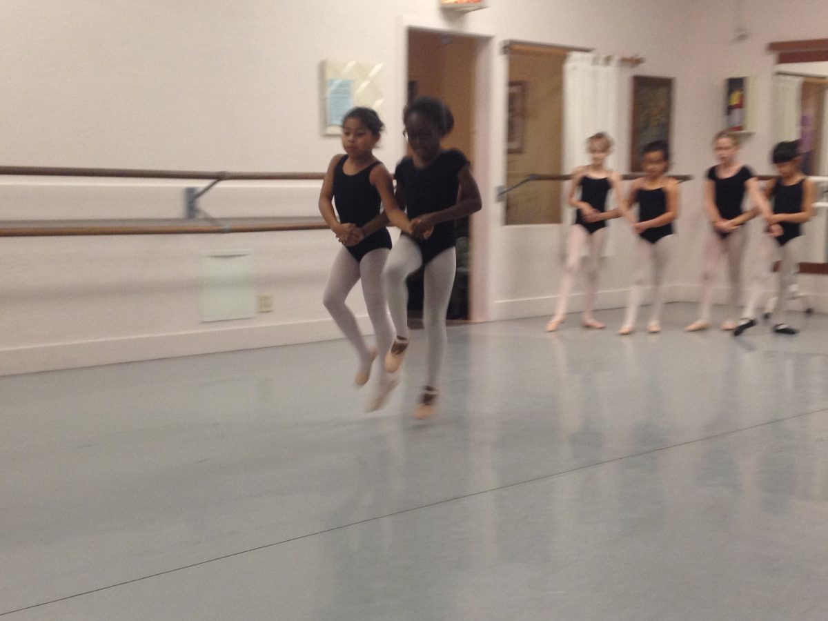 2 young dancers hold hands and skip across the floor together. Other duos wait off to the side for their turn.