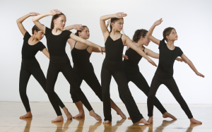 6 dancers in a pose with one hand near their forehead and the other reaching to the side. Dancers are dressed in all black.