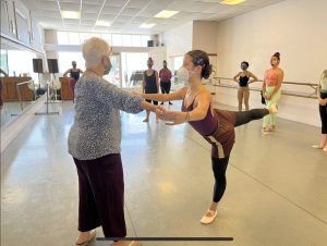 A dancers in a mask stands in arabesque receiving instruction from Beth Hoge, Danspace's founder. Other students in class look on in the background.