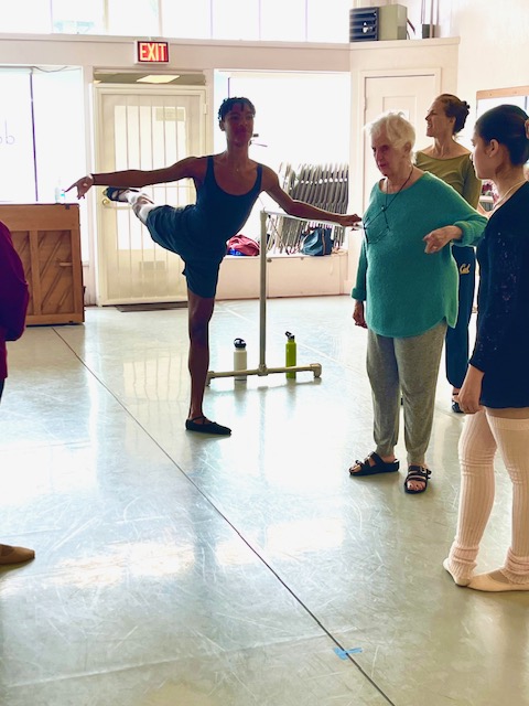 A dancer stands at the barre in arabesque, with other dancers observing and Beth Hoge giving correction.