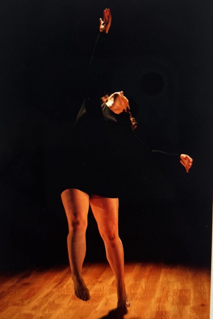 Mary's face is obscured as she reaches up to the ceiling in motion on a wood floor with a dark background.