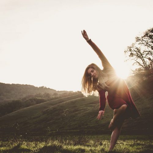 Casey in a red dress and long hair loose poses with one arm up and one leg in passe in front of a green hill with sunlight shining behind her.