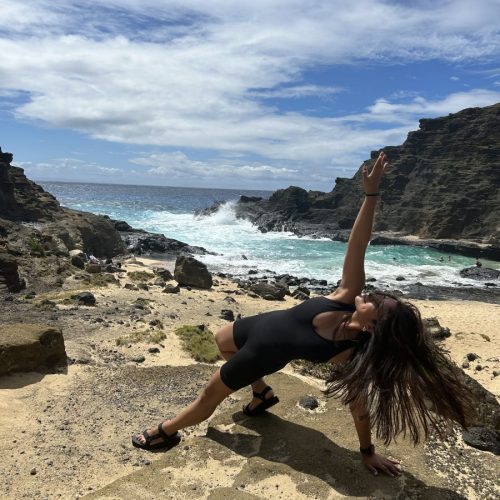 Jocelyn poses with hair in her face, hand reaching up, on a rock in front of a beach of vibrant blue water.