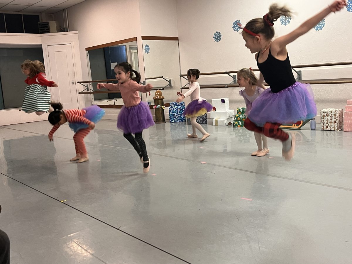 6 young dancers in purple fluffy tutus mid dance in front of a backdrop of prop wrapped gifts