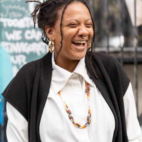 Rivka laughing in front of a colorful background wearing a white shirt, brown shawl, and a colorful necklace.