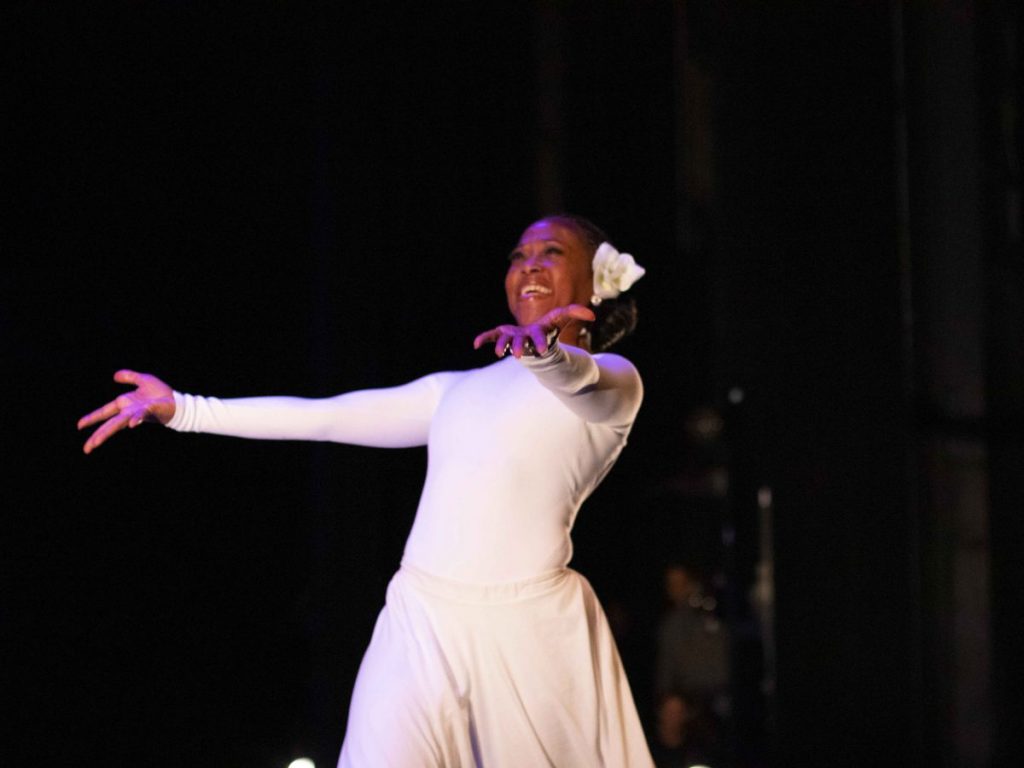 Denice in a white outfit with a white flower in her hair is dancing against a dark backdrop, smiling and reaching forward.