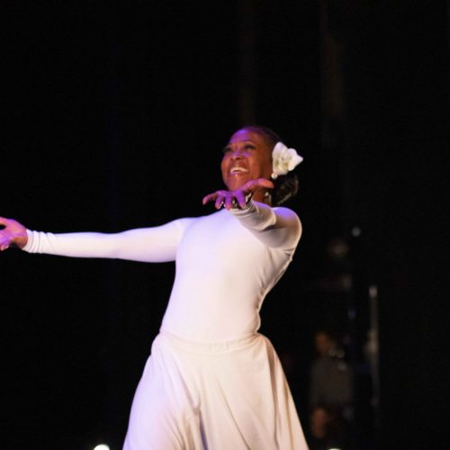 Denice in a white outfit with a white flower in her hair is dancing against a dark backdrop, smiling and reaching forward.