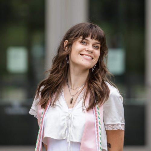 Ellie wears a white and pink top and smiles at the camera. Her brown wavy hair is loose. The background is blurred and Ellie is in focus.